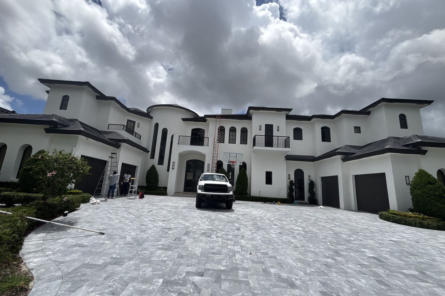 A large white two-story stucco house with a paver driveway, a work truck parked out front and extension ladders set against the facade.