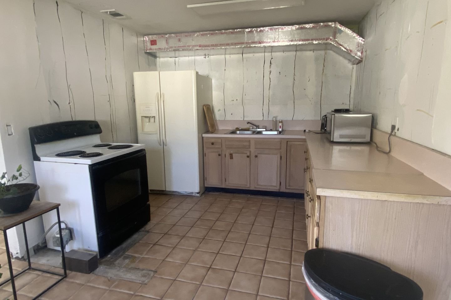 Dated kitchen with pickled oak cabinets, laminate counters, terracotta tile floor and bare walls where the wall cabinets have been removed.
