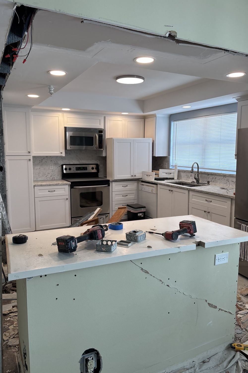 A kitchen part-way through renovation, with a soffit opened to expose the framing, new recessed lights in the ceiling and cordless drills sitting on the island.